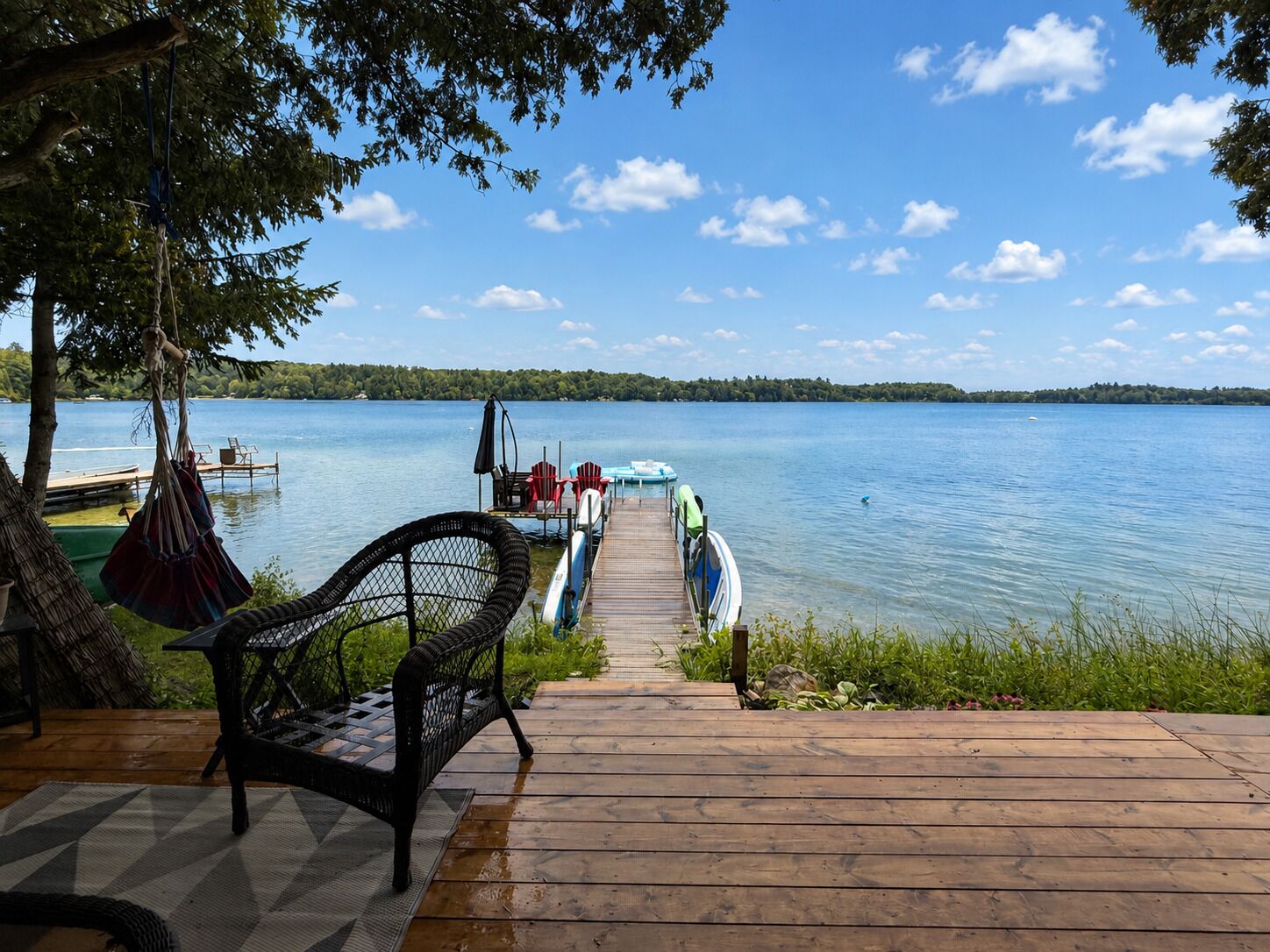 A wooden dock leading to a calm lake with a clear sky, surrounded by greenery and equipped with seating and a small boat.