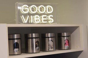 A shelf with a neon sign reading 'GOOD VIBES' and four stainless steel containers with labels.