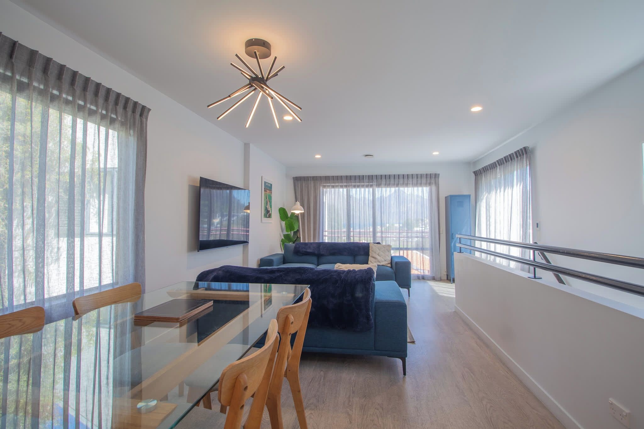 A modern living room with a glass dining table, a blue sofa, and a contemporary ceiling light fixture.
