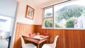 Ein Eckzimmer mit Holztisch, zwei weißen Stühlen, einem Fenster mit Blick auf Häuser und Berge sowie einem gerahmten Bild an der Wand.