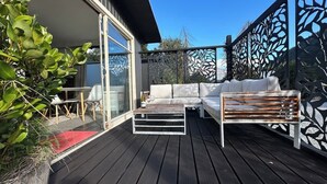 Eine Holzterrasse mit einem weißen Sofa und einer Bank, einer Glastür und einem dekorativen Metallzaun.