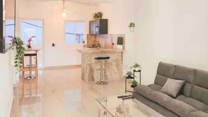 Amplia sala de estar con un sofá gris, una mesa de centro de vidrio y una cocina al fondo.