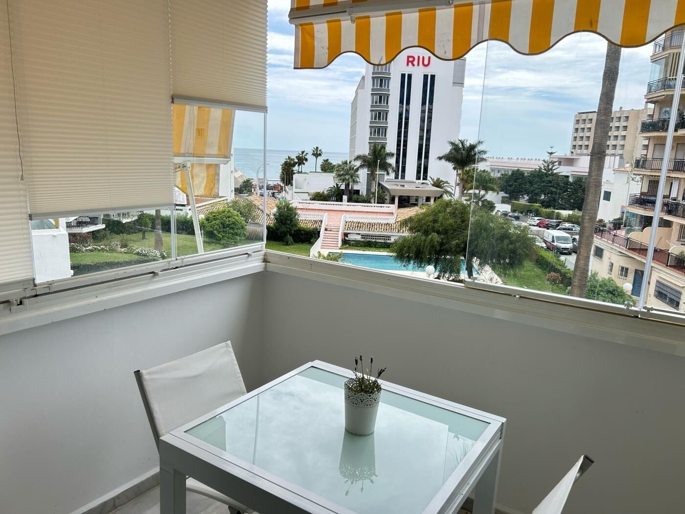 A balcony with a glass table and a potted plant, overlooking a pool and buildings with a prominent RIU sign.