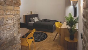 Ein gemütlicher Raum mit Holzboden im Fischgrätmuster, einem Bett mit dunklem Kopfteil, einem kleinen Schreibtisch mit Pflanze und einem gelben Stuhl.