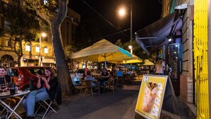 Ein Straßencafé mit Tischen und Stühlen unter Sonnenschirmen, umgeben von Gebäuden und Bäumen.