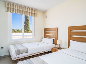 Ein Hotelzimmer mit zwei Betten, einem Fenster mit Blick auf Bäume, einem Nachttisch mit Lampe und einem Wandventilator.