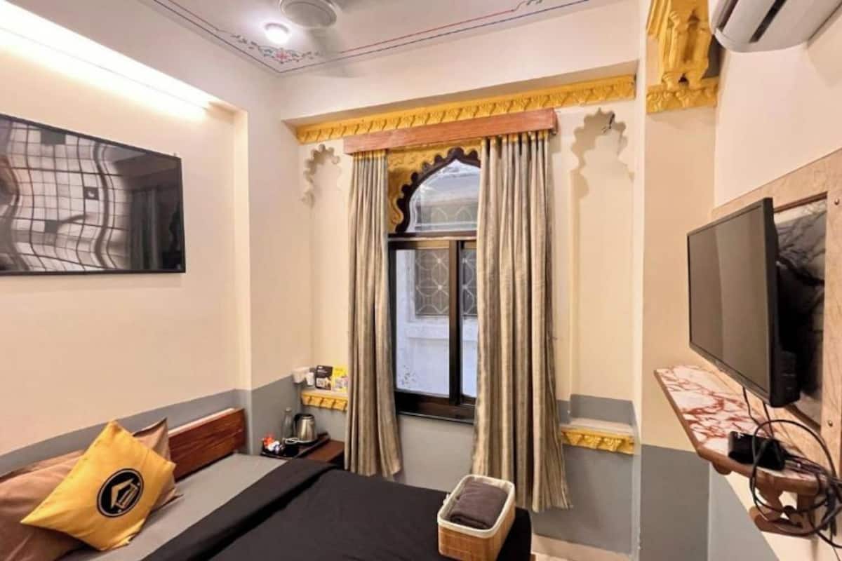 A hotel room with a bed, a television, a window with curtains, a small table with a tray, and a wall-mounted TV.