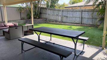 Un patio al aire libre con una mesa de picnic y bancos, rodeado por una cerca de madera y un césped.
