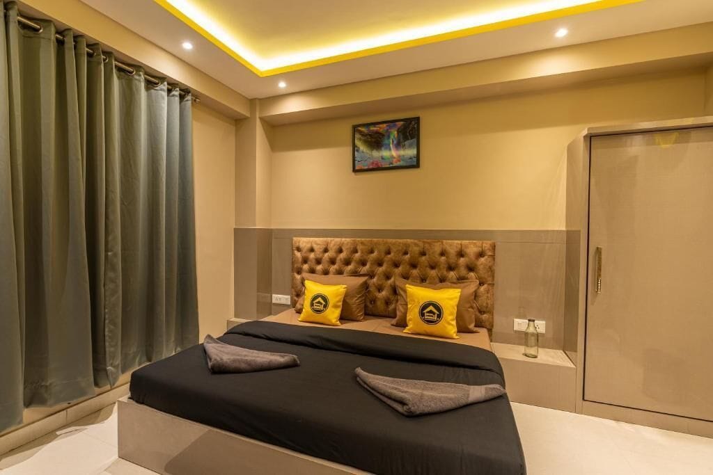 Una habitación de hotel moderna con un cabecero capitonado, una cama grande con ropa de cama negra y gris, dos almohadas amarillas con un logo, un cuadro en la pared y un armario.