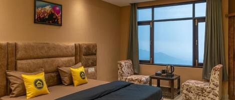 A hotel room with a large bed, two armchairs, a small table, and a large window offering a mountain view.
