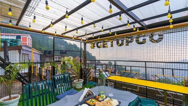 An outdoor dining area with a table set for two, surrounded by green chairs, under a glass roof with hanging lights. - The Hosteller Shimla (Shimla)