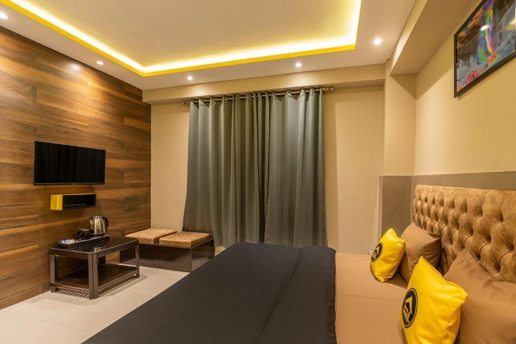 A modern bedroom with a large bed, a flat-screen TV mounted on a wooden wall, a small table with a kettle, and a window with curtains.