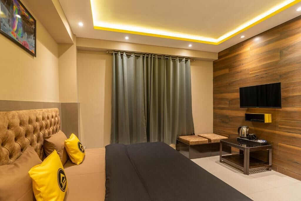 Ein modernes Hotelzimmer mit einem gepolsterten Kopfteil, einem Flachbildfernseher an einer Holzwand, einem kleinen Tisch mit Wasserkocher und einem großen Fenster mit Vorhängen.