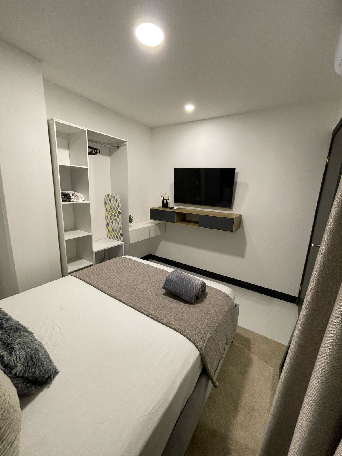 Et moderne soveværelse med seng, fjernsyn ophængt på væggen, skab og en lille hylde med lampe.