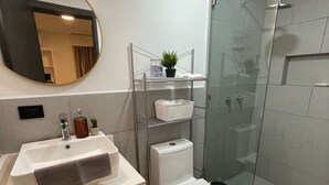 Een moderne badkamer met een wit wastafelmeubel, spiegel en toilet. Er is een douchecabine van glas en een houten kastje.