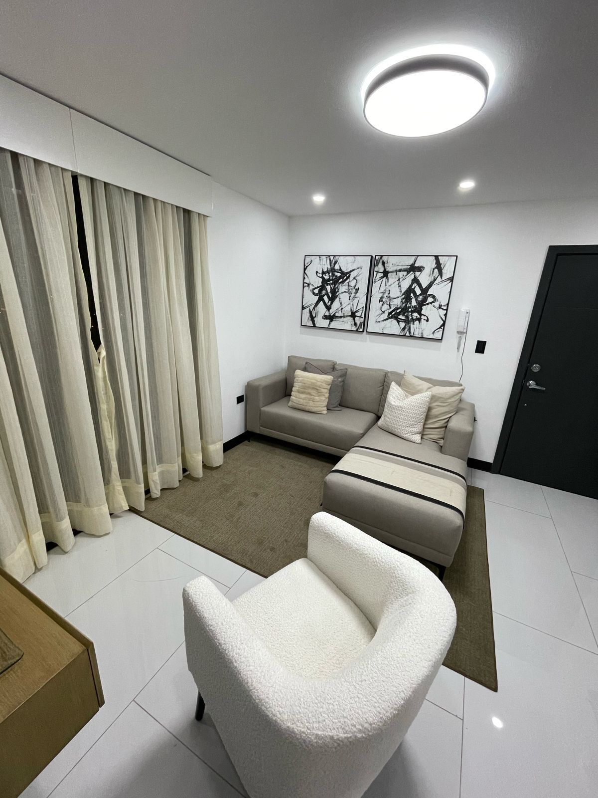 A modern living room with a sofa, a white armchair, and a coffee table. There are abstract paintings on the wall and a door in the background.