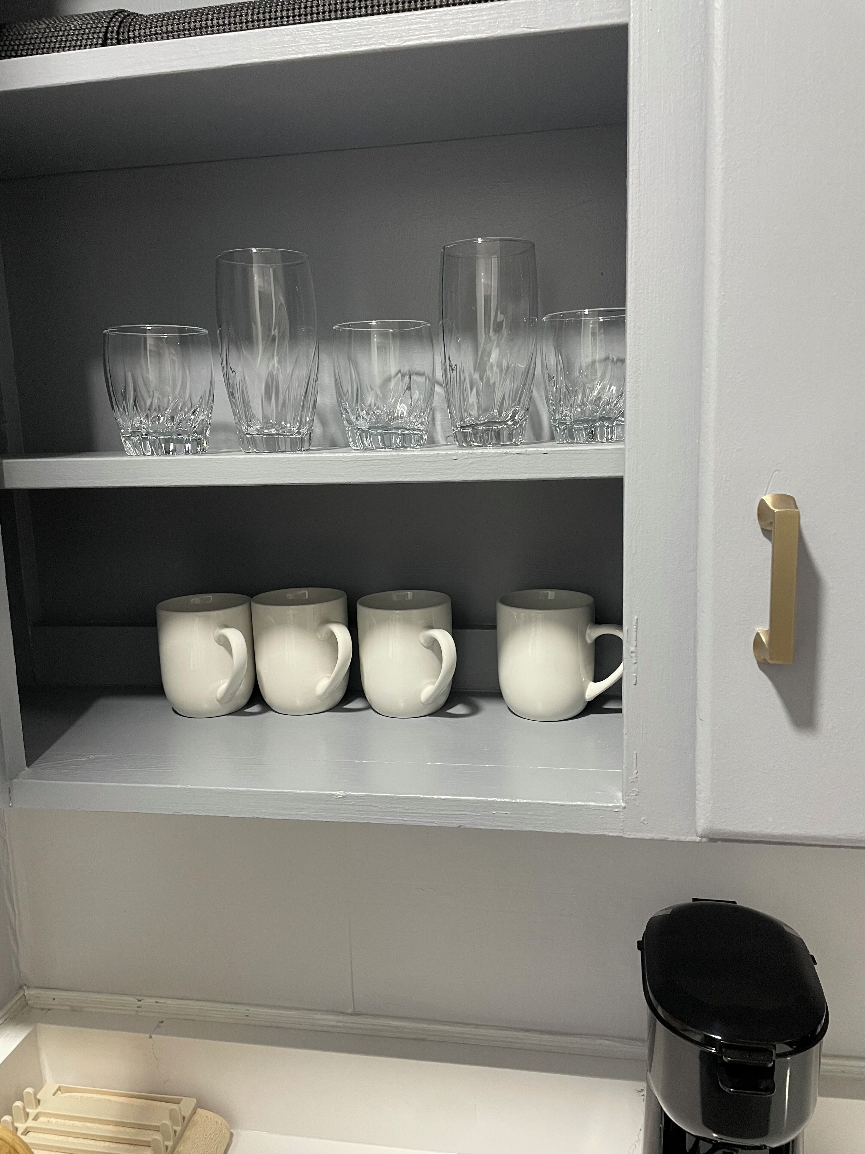 A kitchen with white cabinets, a coffee maker, and a shelf with clear glasses and white mugs.