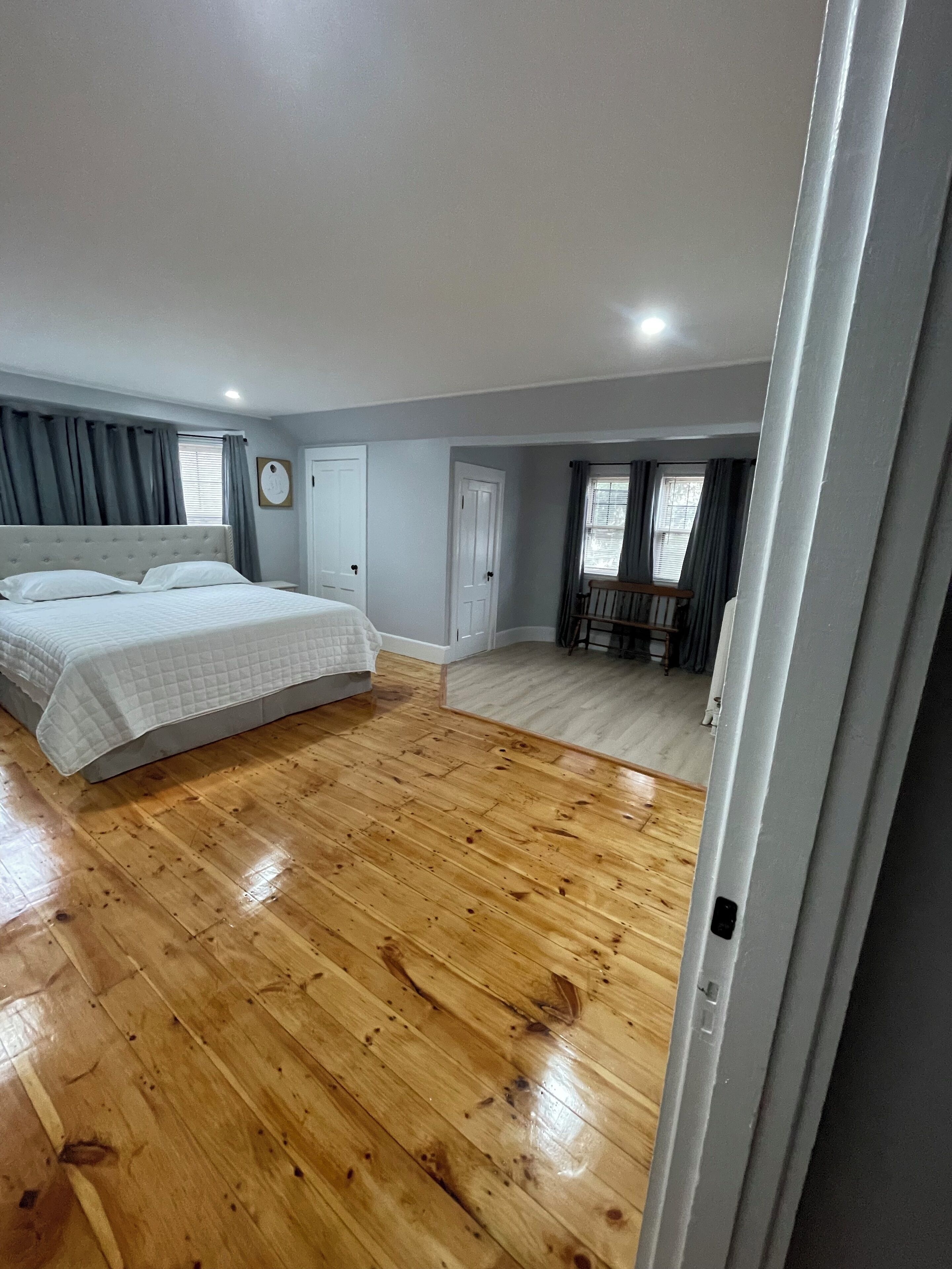 Un dormitorio con piso de madera, una cama con edredón blanco, una silla de madera y una ventana con cortinas.