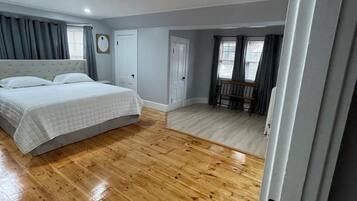 Un dormitorio con piso de madera, una cama con edredón blanco, una silla de madera y una ventana con cortinas.