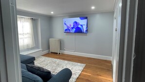 Una sala de estar con un televisor en la pared, un sofá azul, un radiador y una ventana con cortinas.