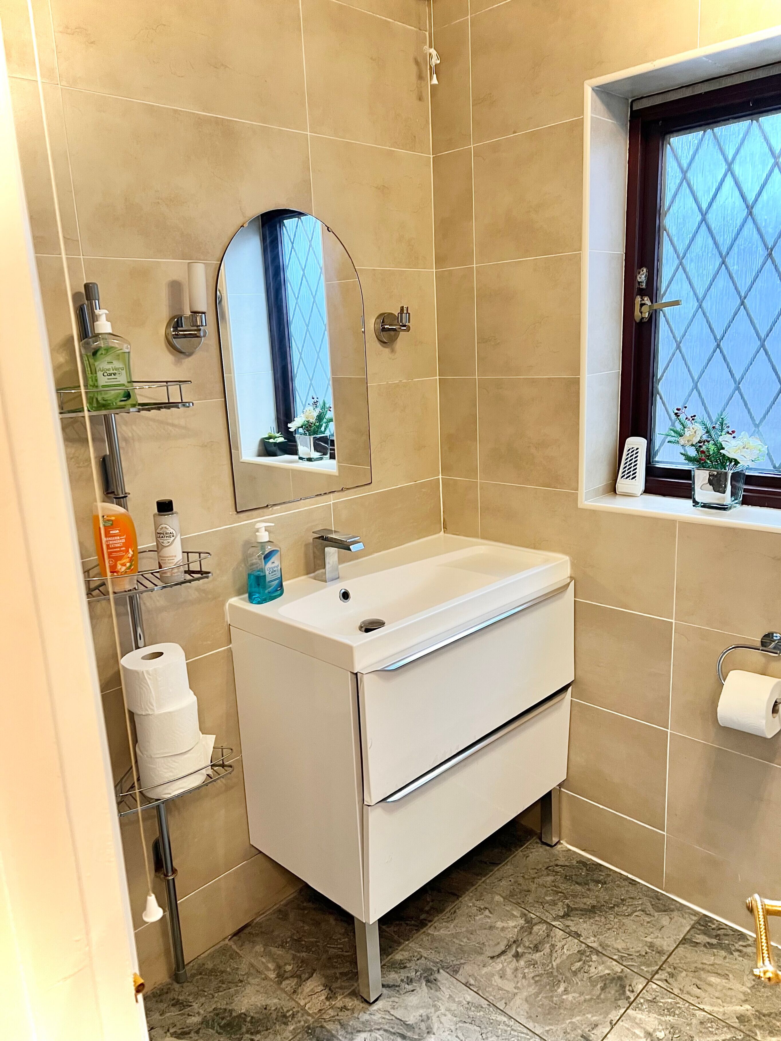 Une salle de bain avec un lavabo blanc, un miroir, une fenêtre à motifs losangés et une étagère contenant des articles de toilette.