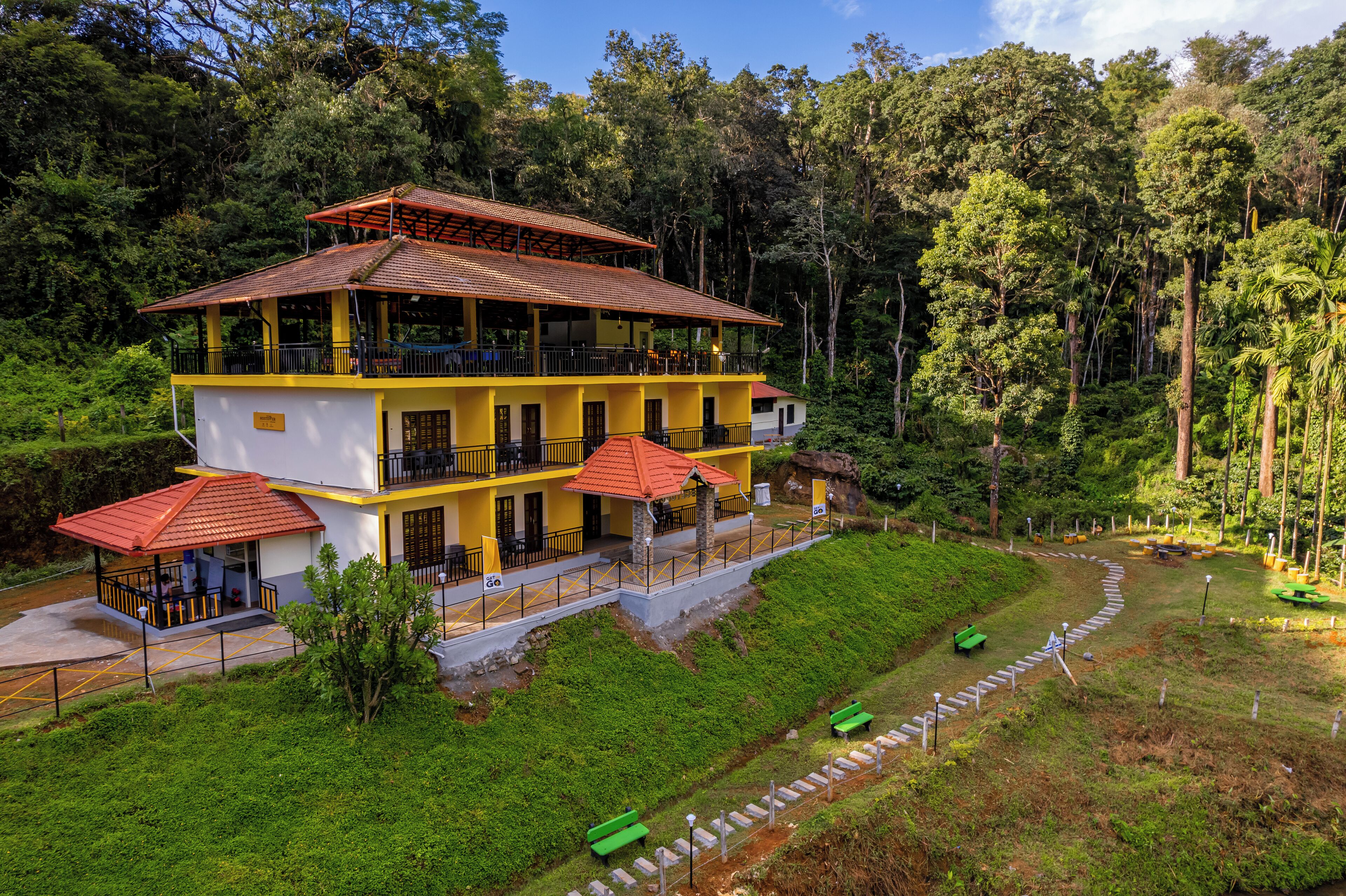 A multi-story yellow building with balconies, surrounded by lush greenery and a forested area.
