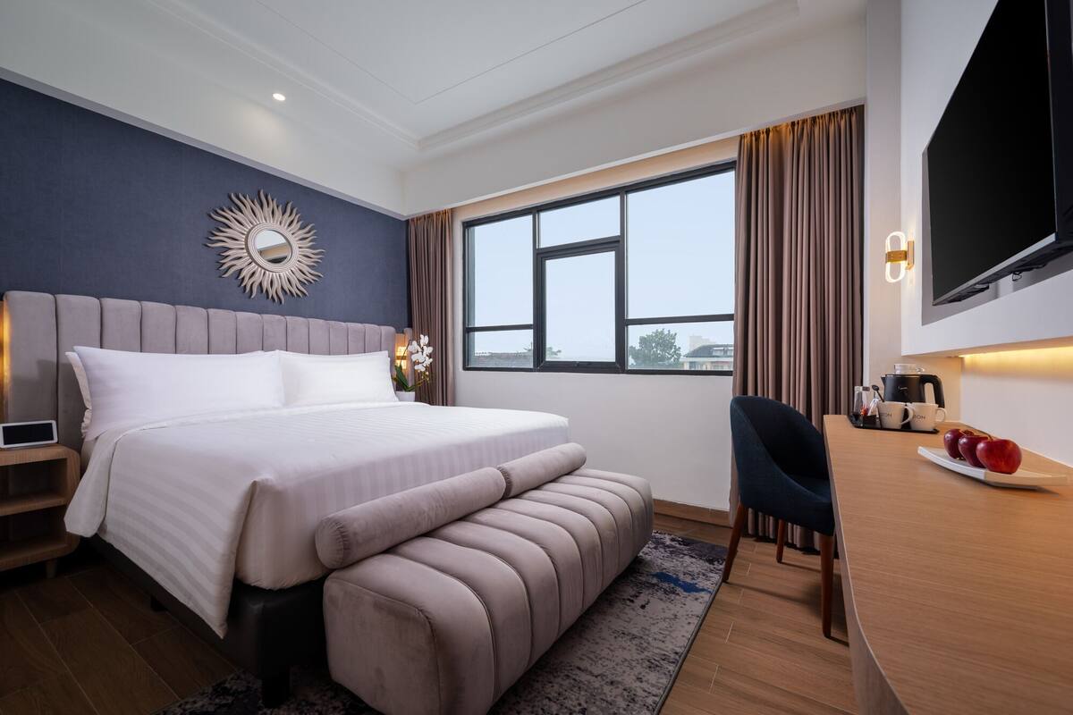 A modern hotel room with a large bed, a desk with a chair, a flat-screen TV, and a window with curtains.