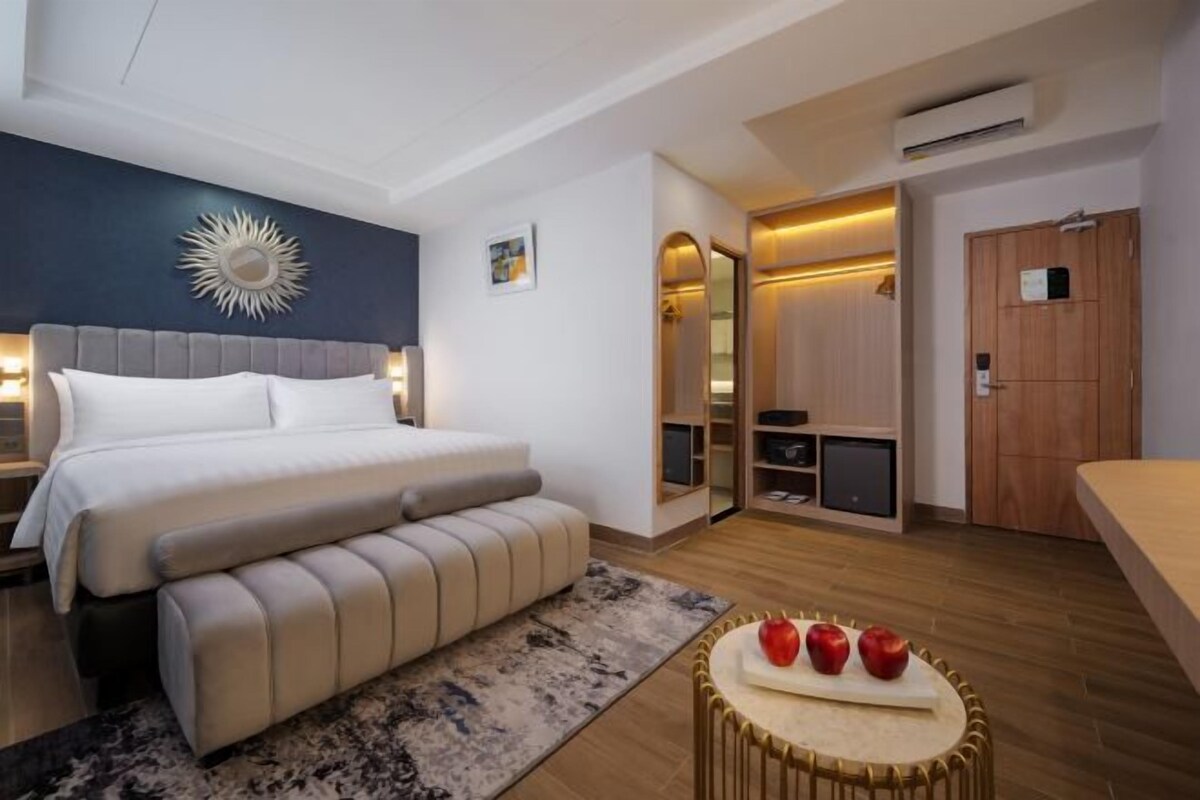 A modern bedroom with a large bed, bedside tables, a bench, a mirror, a wardrobe, and a small table with apples.