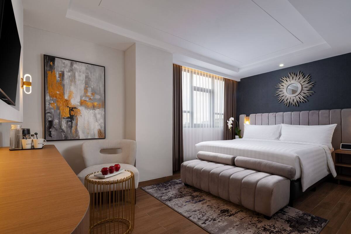 A modern hotel room with a large bed, a bedside table, a chair, and a small round table with apples.