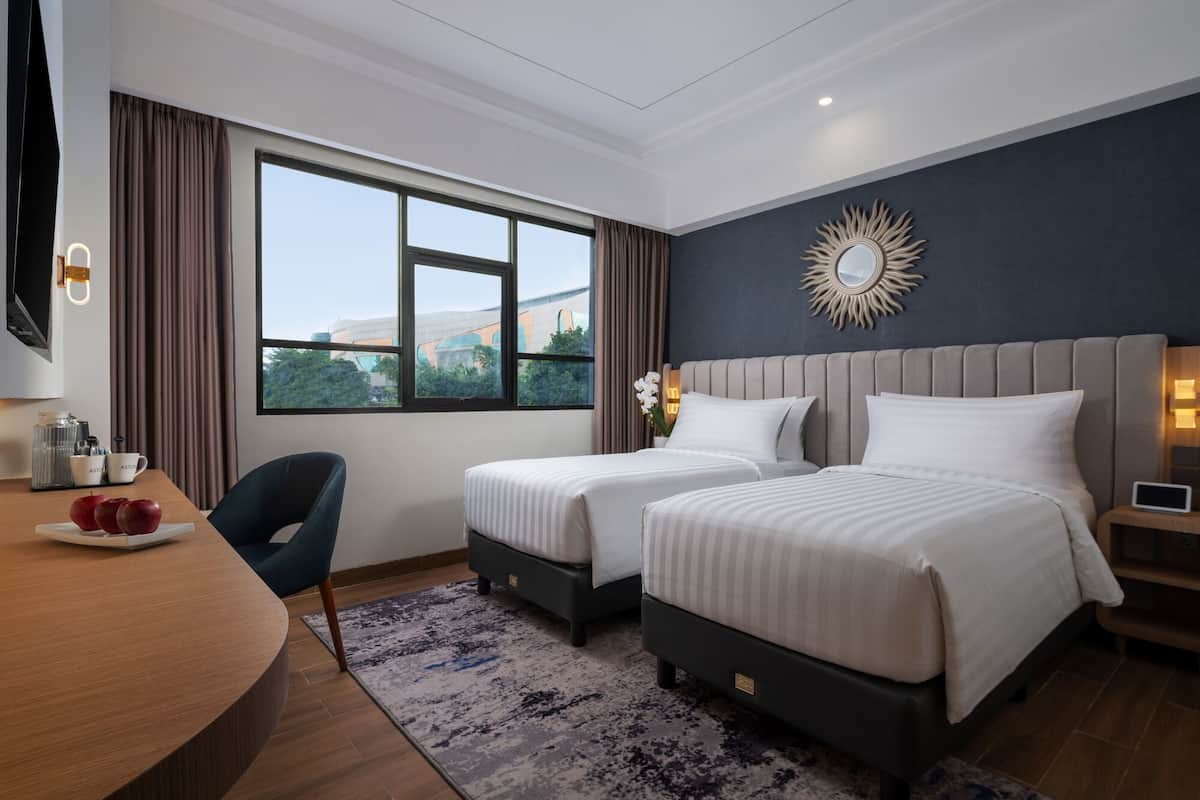 A hotel room with two beds, a desk, a chair, and a large window offering a city view.