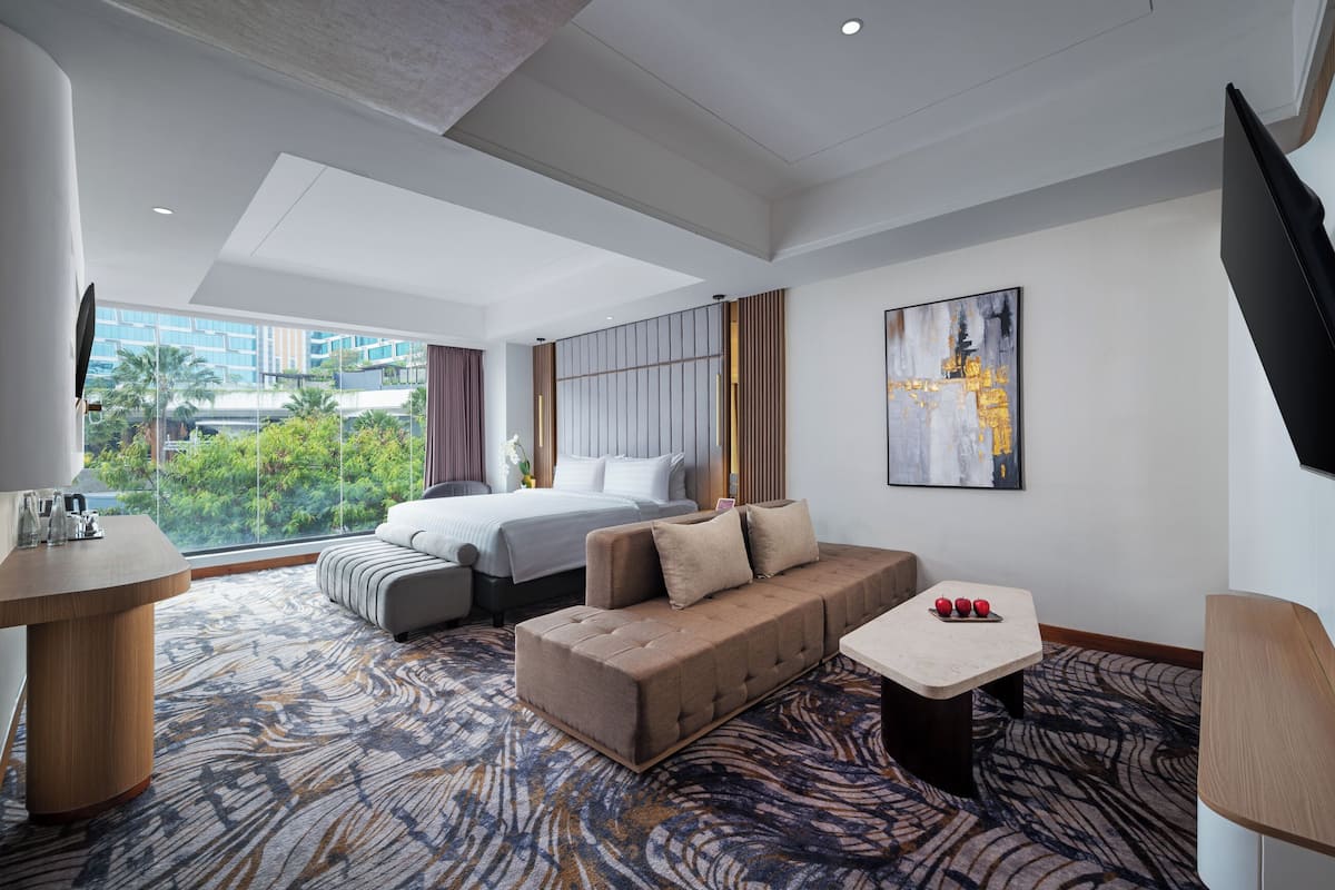A modern hotel room with a large bed, a sofa, a small table with apples, a TV, and a view of greenery outside.