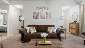 A living room with a brown sofa, a coffee table, and a vase of white flowers. There are three framed artworks on the wall above the sofa. - Relax, Quiet, Clean, & 15min Away From Airport (Houston)