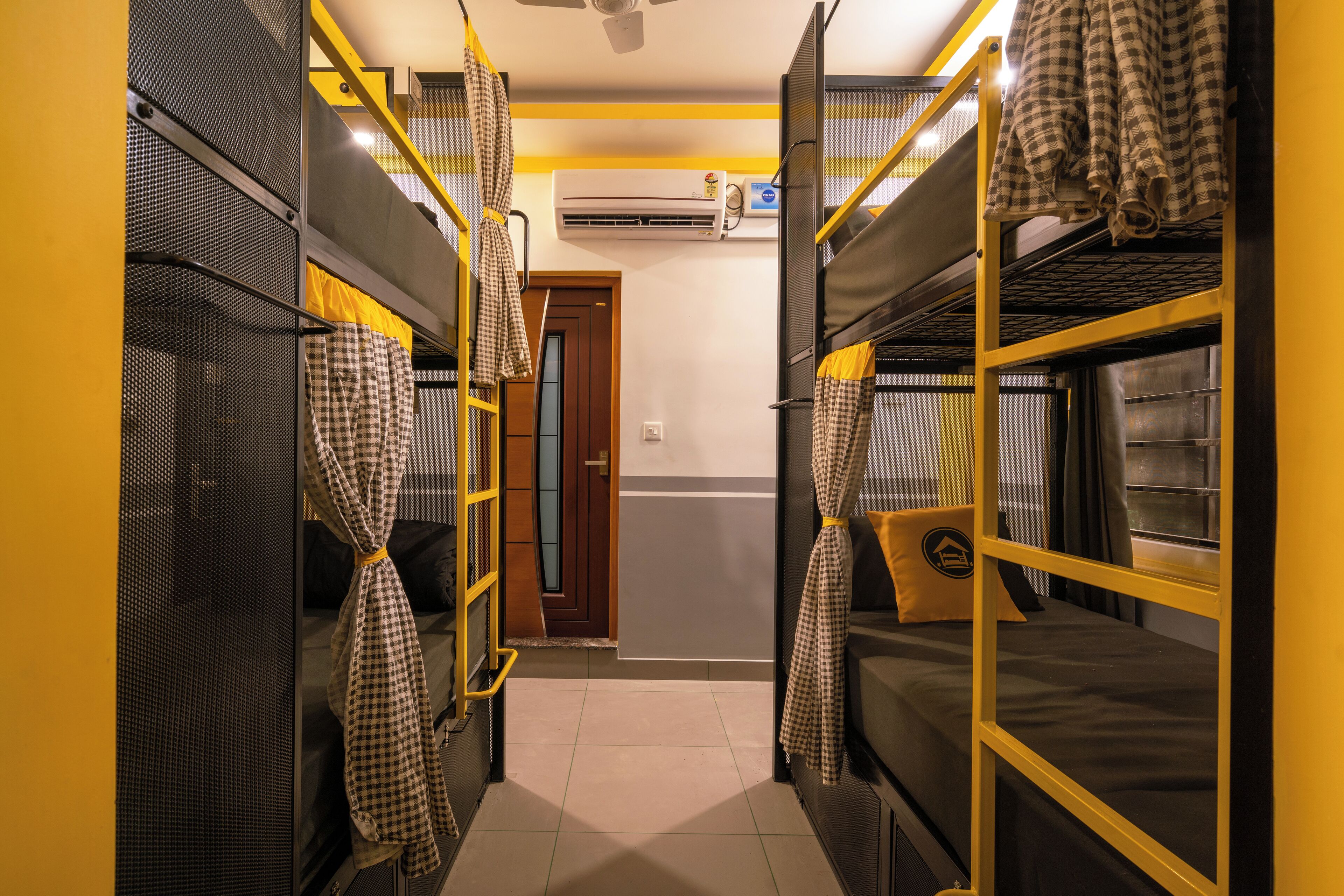 A dormitory room with bunk beds, a door, an air conditioning unit, and a window with curtains.