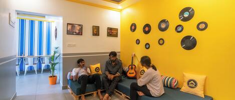 Une pièce avec un mur jaune, une guitare et un groupe de personnes assises sur un banc.