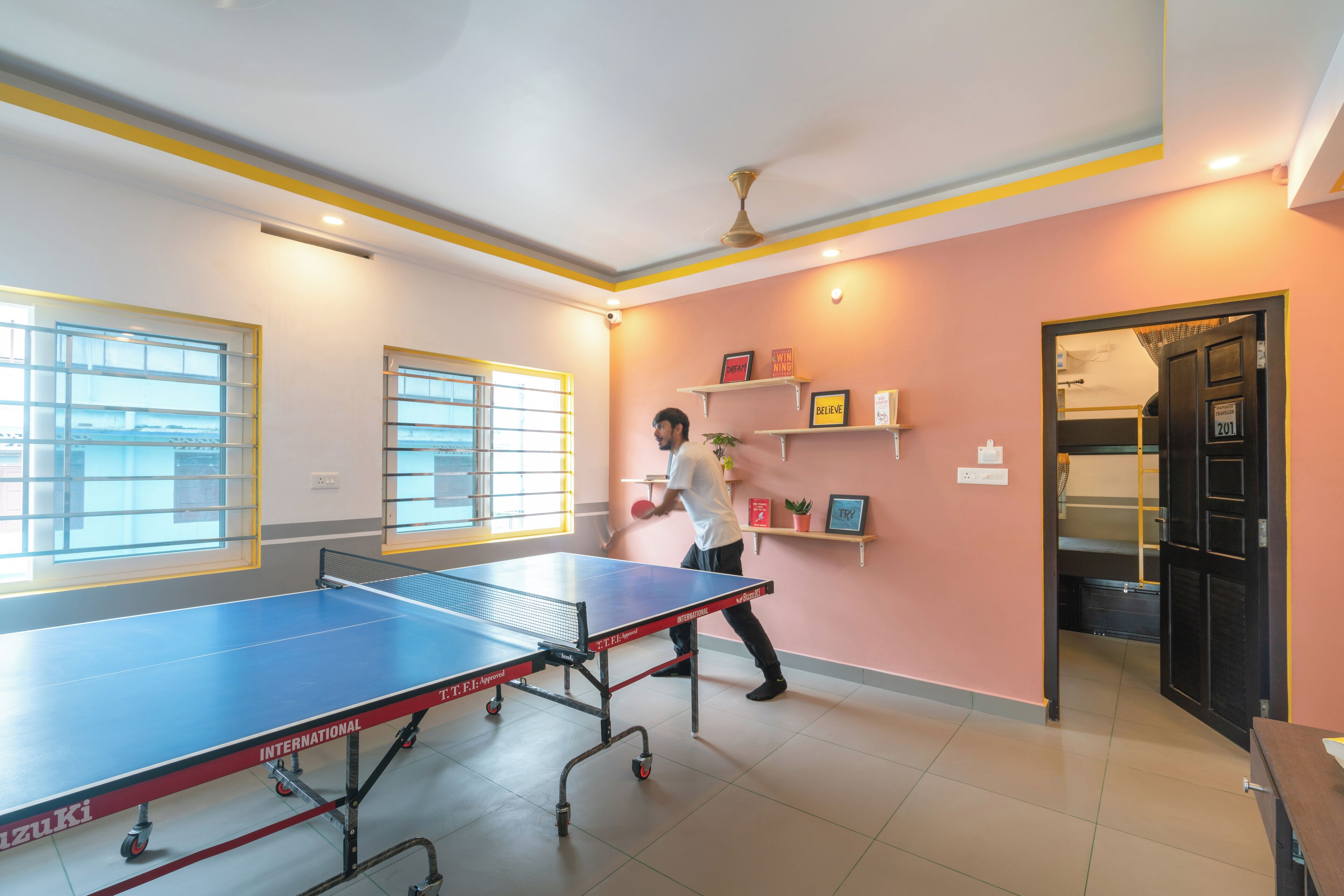 Une table de ping-pong dans une pièce aux murs roses, avec un ventilateur au plafond et des étagères ornées de citations inspirantes.