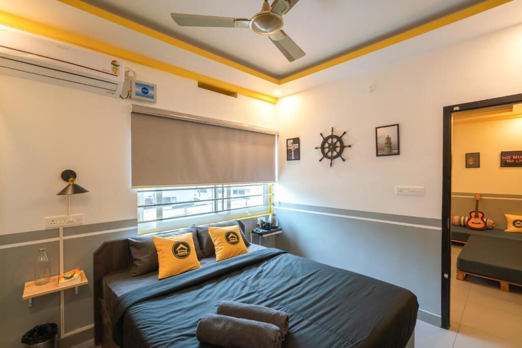 A hotel room with a bed, a ceiling fan, a window with blinds, a wall clock, and a small table with a lamp.