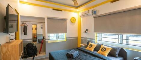 Habitación de hotel con cama, escritorio, espejo, ventilador de techo y ventana con persianas.