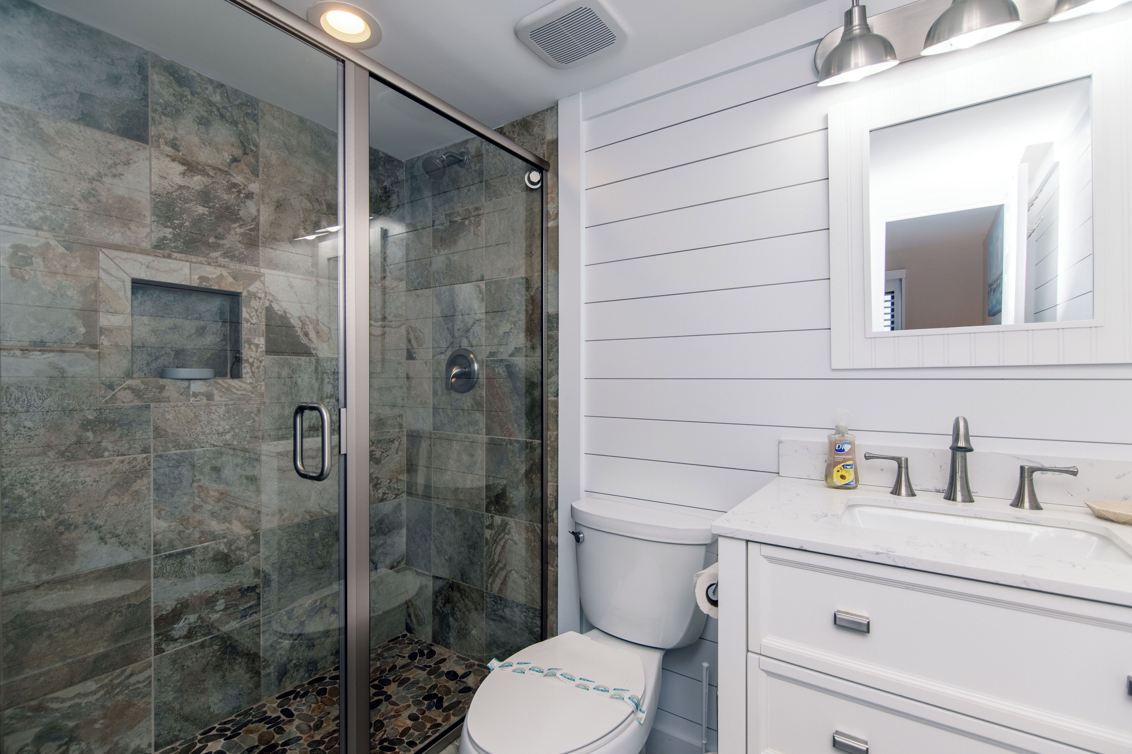 Une salle de bain avec une douche à parois de verre, des toilettes blanches, un lavabo avec un miroir au-dessus et un petit flacon de savon posé sur le comptoir.