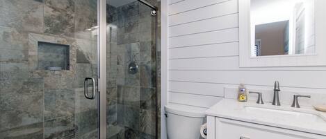 Une salle de bain avec une douche à parois de verre, des toilettes blanches, un lavabo avec un miroir au-dessus et un petit flacon de savon posé sur le comptoir.