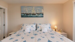 Un dormitorio con una cama con colcha azul estampada con motivos florales, un cuadro de veleros en la pared y una mesita de noche con una lámpara y una caja de pañuelos.