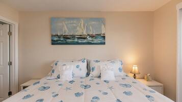 A bedroom with a bed featuring a blue floral bedspread, a painting of sailboats on the wall, and a bedside table with a lamp and a tissue box.