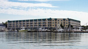 Un hotel frente al mar con varios balcones, un puerto deportivo con barcos y un cielo despejado.