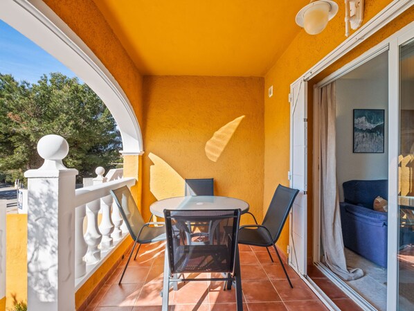 A balcony with a round table and chairs, a white railing, and a sliding glass door leading to an interior room with a blue sofa.