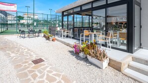 Un patio exterior con mesas y sillas, un área de comedor con paredes de vidrio y vistas al paisaje circundante.