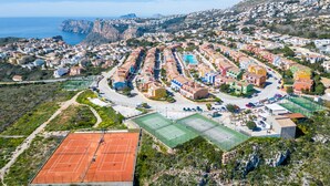 Vista aérea de un pueblo costero con cancha de tenis, edificios residenciales y montañas al fondo.