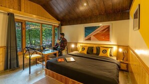 Ein gemütliches Zimmer mit Holzbalkendecke, einem Bett mit schwarzer Bettwäsche, einem Schreibtisch mit Stuhl und einem großen Fenster mit Vorhängen.