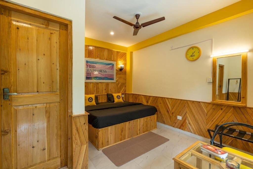 A room with a bed, a wooden door, a ceiling fan, a clock, and a mirror.