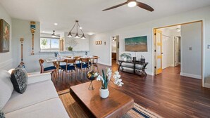 A spacious living room with a dining area, wooden flooring, ceiling fan, and a painting of a beach scene. - 2BD Pool & Resort's Amenities, Ocean Views! - Kaanapali Plantation 34 (Lahaina)