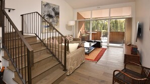 A spacious living area with a wooden staircase, a sofa, a coffee table, and a large window offering a view of the outdoors.