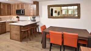 A kitchen with wooden cabinets, a stainless steel microwave, a sink, and a bar area with stools. There is a dining table with orange chairs and a large mirror above it.